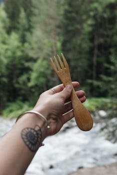 Tattooed hand holding a wooden fork with a forest background, showcasing eco-friendly utensils.